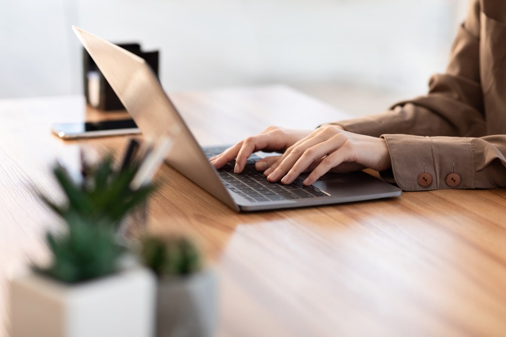 A woman is typing on her laptop with plants on the desk next to her.
